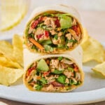 Two halves of a chicken salad wrap, with veggies stacked on top of each other on a plate, along with potato chips.