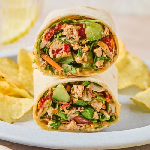 Two halves of a chicken salad wrap, with veggies stacked on top of each other on a plate, along with potato chips.