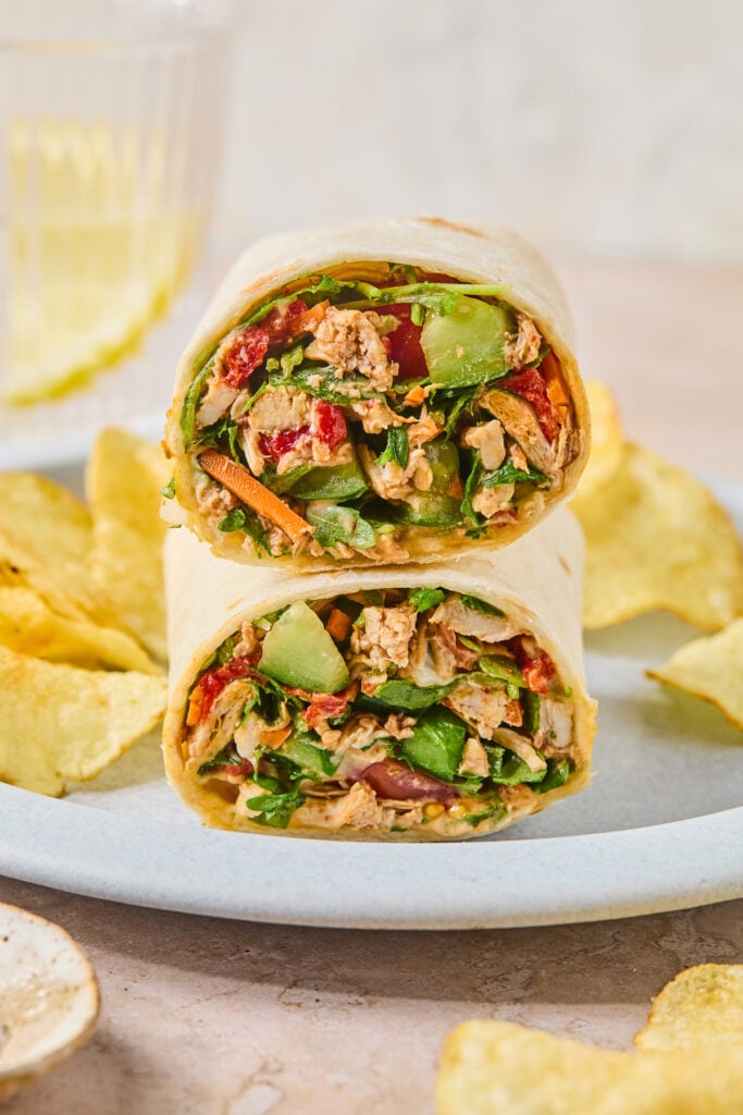 Two halves of a chicken salad wrap, with veggies stacked on top of each other on a plate, along with potato chips.