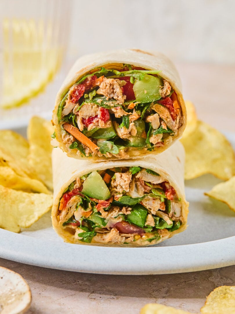 Two halves of a chicken salad wrap, with veggies stacked on top of each other on a plate, along with potato chips.