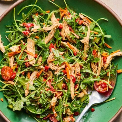 A shallow green bowl filled with a mix of arugula, shredded carrots, chopped cucumbers, halved cherry tomatoes, shredded chicken, and goat cheese tossed in olive oil and vinegar. There is also a fork in the bowl. Next to the bowl is a small bowl of salt and pepper.