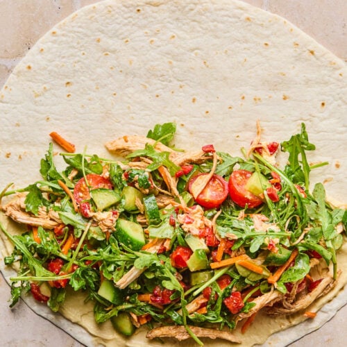 A large tortilla lay flat with hummus and a salad mixture with chicken spread across the bottom half.
