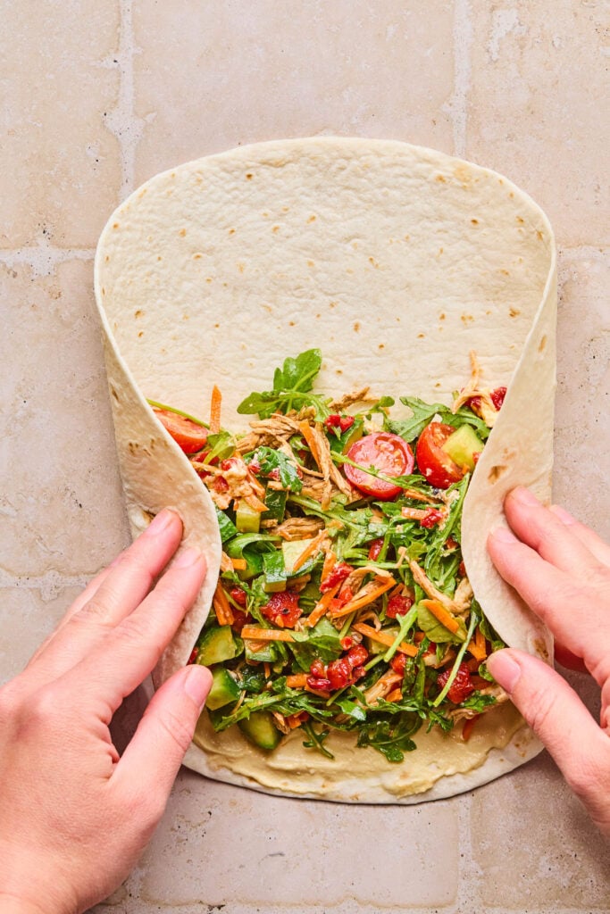 A large tortilla lay flat with a salad mixture spread across the bottom half. The left and right sides are being folded to wrap it up.