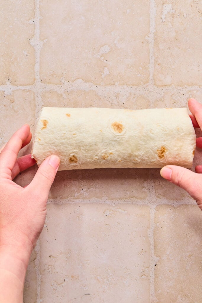 Hands holding a chicken salad sandwich wrap on a tan tile counter. It has just been wrapped up.
