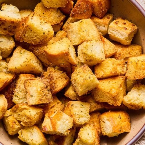 A close up of a bowl of sourdough croutons. Next to the bowl is a tan linen towel.