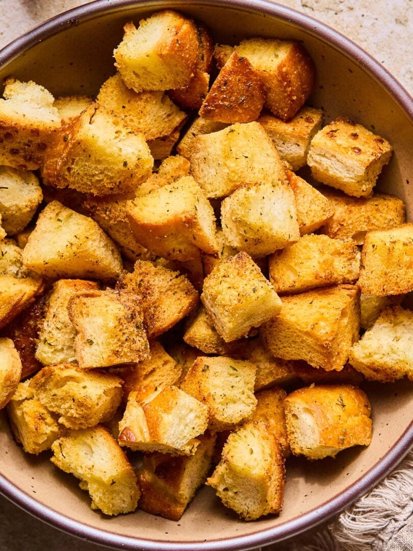 A close up of a bowl of sourdough croutons. Next to the bowl is a tan linen towel.