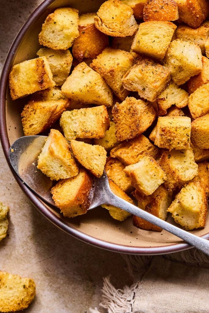 Sourdough croutons in a bowl with a metal spoon. Around the bowl is a tan linen towel and some croutons.