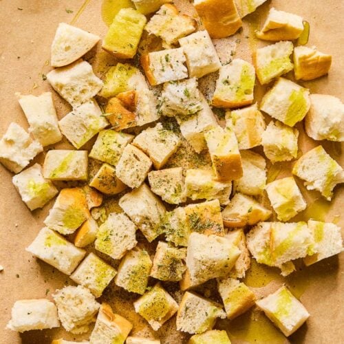 Cubed bread on a baking sheet lined with parchment paper. It is drizzled with olive oil and spices before it is mixed together.