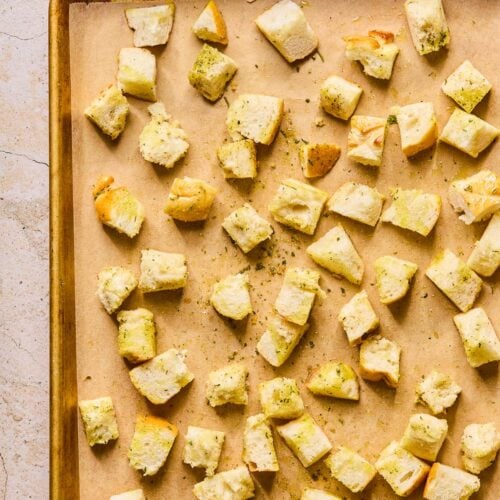 Cubed bread that has been tossed in oil and spices spread out on a parchment paper lined baking sheet. Next to the baking sheet is a cruet of olive oil.