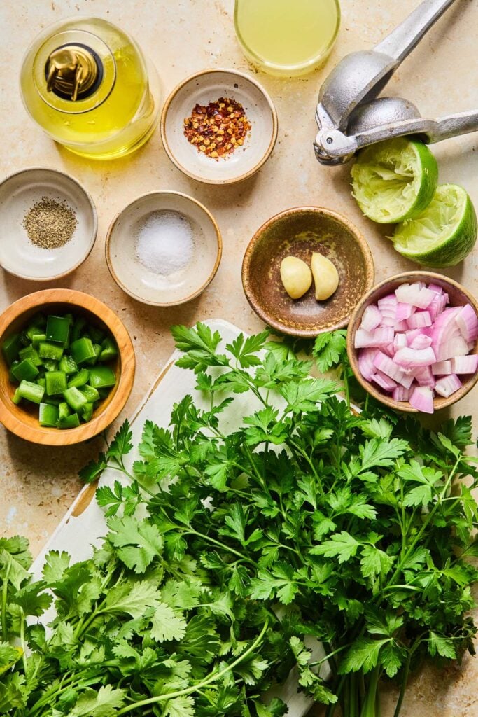Small bowl of lime juice, a citrus squeezer, a lime that has been cut in half and juiced, a small bowl with two cloves of garlic in it, a small bowl of chopped shallot, a bunch of fresh parsley, a bunch of fresh cilantro, a small bowl of chopped jalape&ntilde;o, a small bowl of salt, a small bowl of black pepper, a small bowl of red pepper flakes, and a jar of olive oil on a tan cement counter.