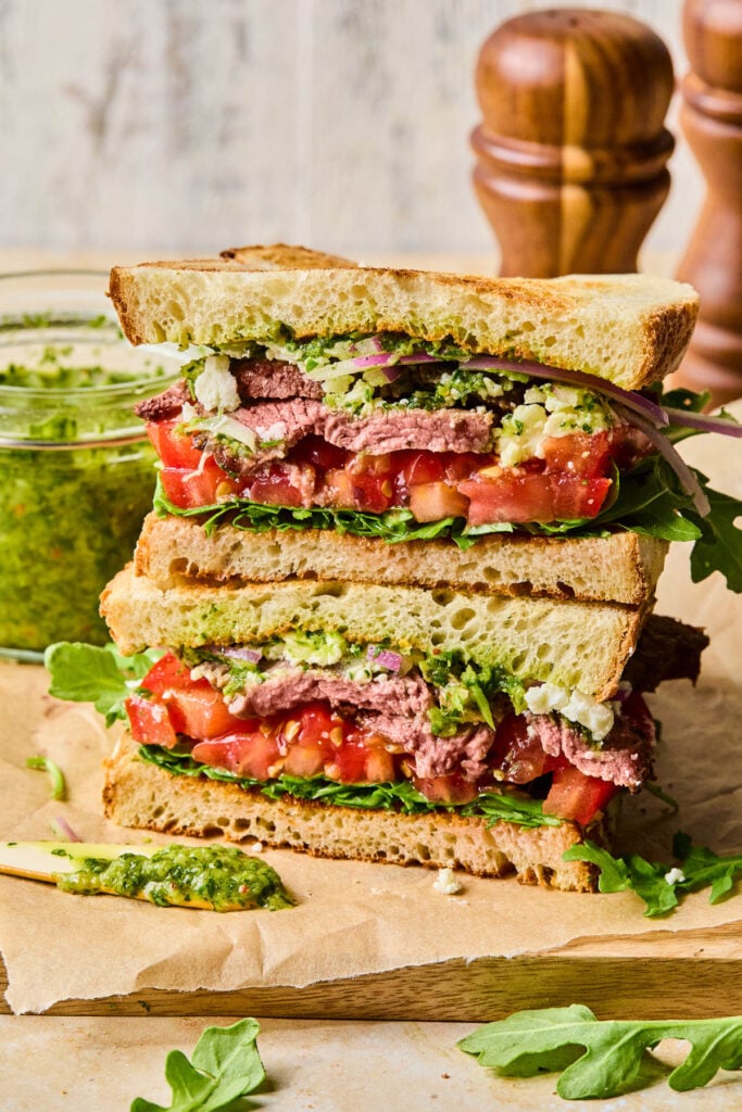 Two halves of a steak sandwich with chimichurri, red onion, goat cheese, tomato, and arugula stacked on top of each other on a cutting board. Behind the sandwich is a jar of chimichurri and some wooden salt and pepper shakers.