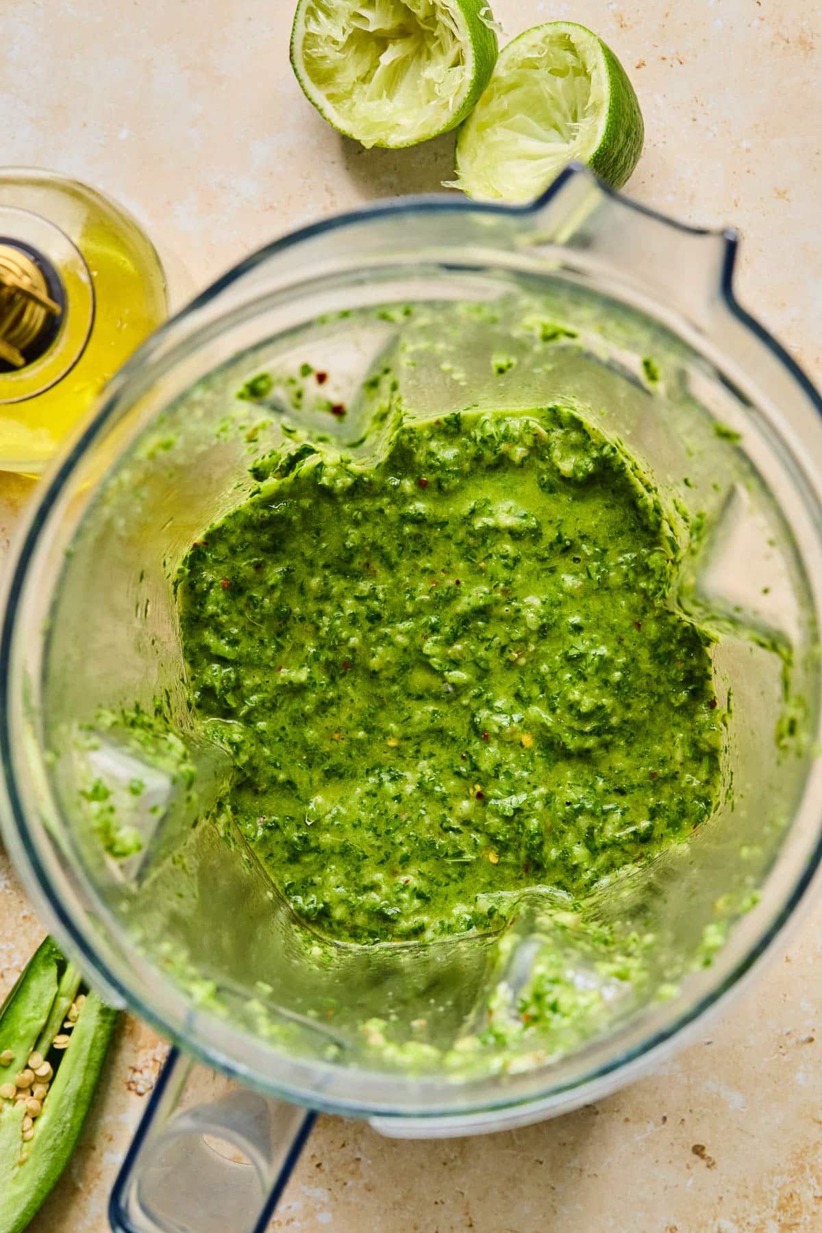 Blender chimichurri sauce in a blender. Around the blender is half a jalape&ntilde;o, a jar of olive oil, and a juiced lime.