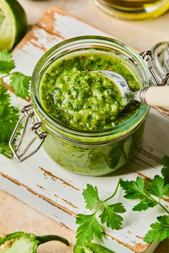Jalape&ntilde;o chimichurri in a jar with a spoon in it. The jar is on a white cutting board with fresh parsley leaves. Around the cutting board are a jalape&ntilde;o, half a juiced lime, and a jar of olive oil.