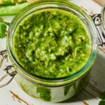 A jar of spicy chimichurri sauce on a white wooden cutting board. Around the jar are some parsley leaves and a jalape&ntilde;o that was cut in half lengthwise.