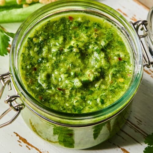 A jar of spicy chimichurri sauce on a white wooden cutting board. Around the jar are some parsley leaves and a jalapeño that was cut in half lengthwise.