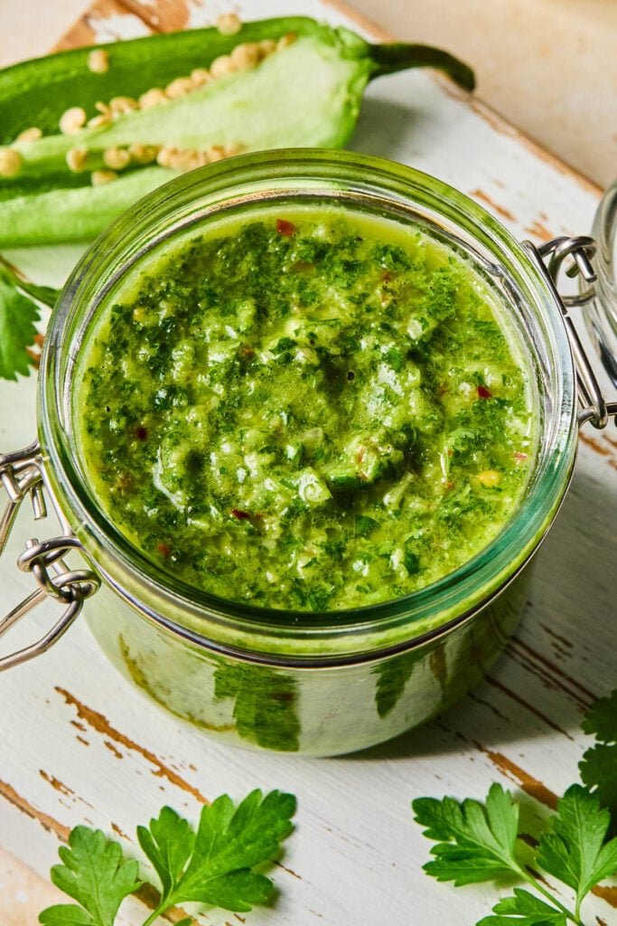 A jar of spicy chimichurri sauce on a white wooden cutting board. Around the jar are some parsley leaves and a jalape&ntilde;o that was cut in half lengthwise.