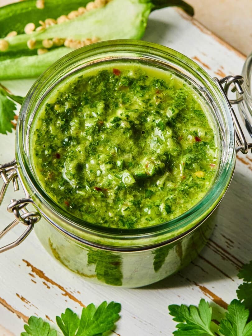 A jar of spicy chimichurri sauce on a white wooden cutting board. Around the jar are some parsley leaves and a jalape&ntilde;o that was cut in half lengthwise.