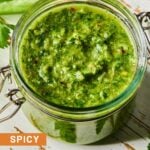 A jar of spicy chimichurri sauce on a white wooden cutting board. Around the jar are some parsley leaves and a jalape&ntilde;o that was cut in half lengthwise.