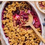 Overhead shot of berry crisp in an oval casserole dish with a wooden spoon in it. A scoop has already been taken out of the crisp. Around the dish is a pink kitchen towel, some fresh berries, and a plate of berry crisp.