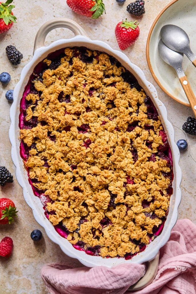 Berry fruit crisp in an oval casserole dish. It is surrounded by some fresh berries, a pink kitchen towel, and a small bowl with two spoons in it.
