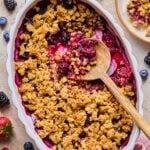 Overhead shot of berry crisp in an oval casserole dish with a wooden spoon in it. A scoop has already been taken out of the crisp. Around the dish is a pink kitchen towel, some fresh berries, and a plate of berry crisp.