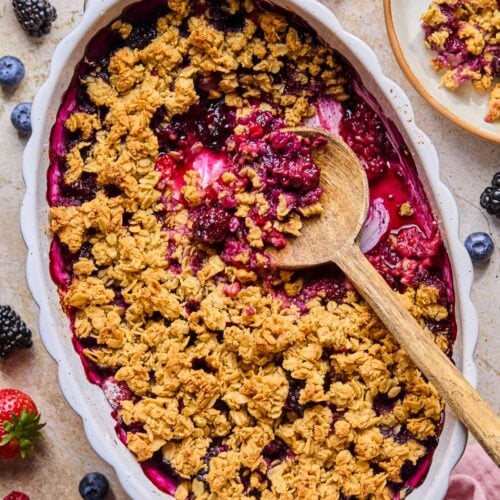 Overhead shot of berry crisp in an oval casserole dish with a wooden spoon in it. A scoop has already been taken out of the crisp. Around the dish is a pink kitchen towel, some fresh berries, and a plate of berry crisp.