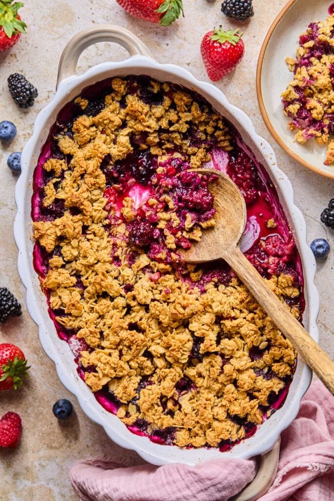 Overhead shot of berry crisp in an oval casserole dish with a wooden spoon in it. A scoop has already been taken out of the crisp. Around the dish is a pink kitchen towel, some fresh berries, and a plate of berry crisp.