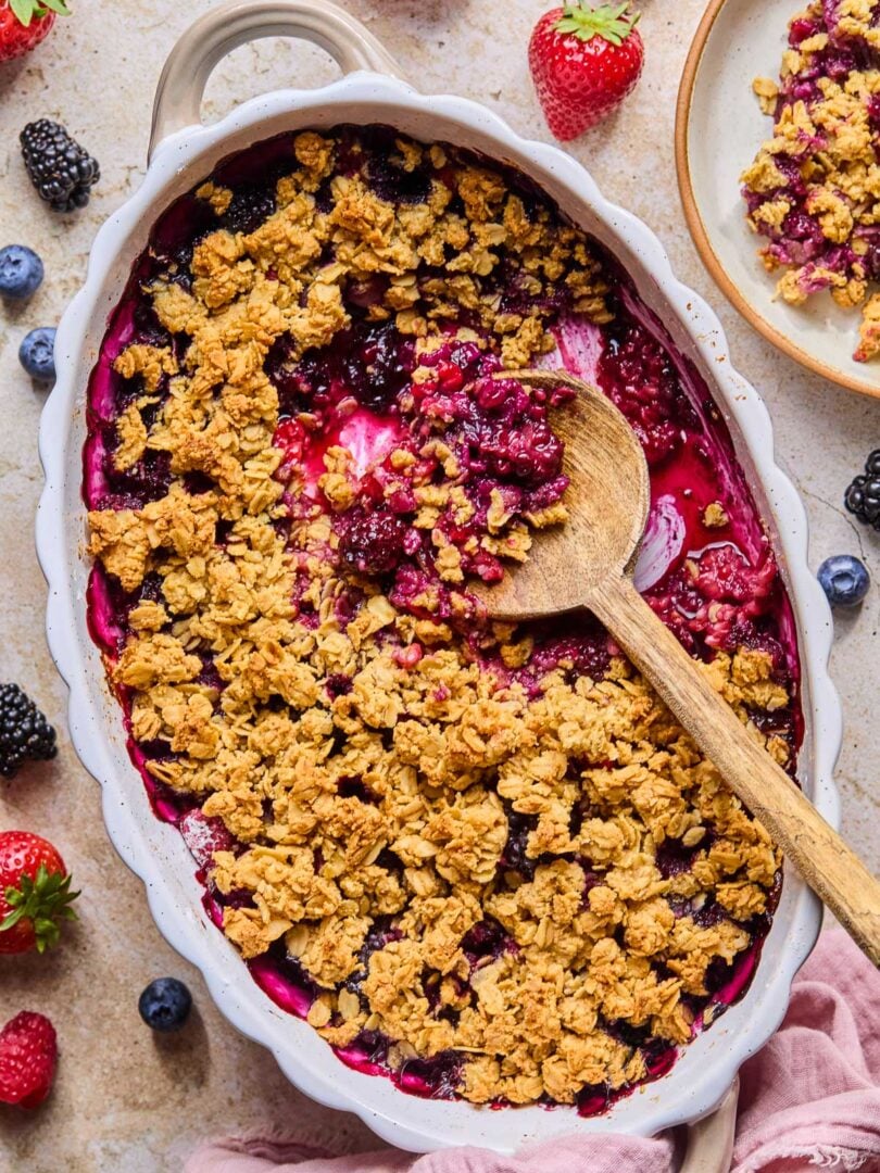 Overhead shot of berry crisp in an oval casserole dish with a wooden spoon in it. A scoop has already been taken out of the crisp. Around the dish is a pink kitchen towel, some fresh berries, and a plate of berry crisp.