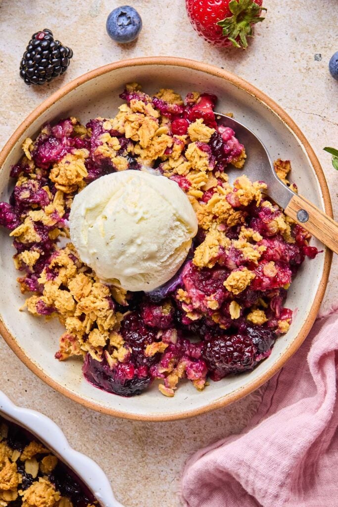 Gluten free berry crisp topped with a scoop of vanilla ice cream in a bowl with a spoon in it. Around the bowl are some fresh blueberries and strawberries, a pink kitchen towel, and the dish of the crisp.