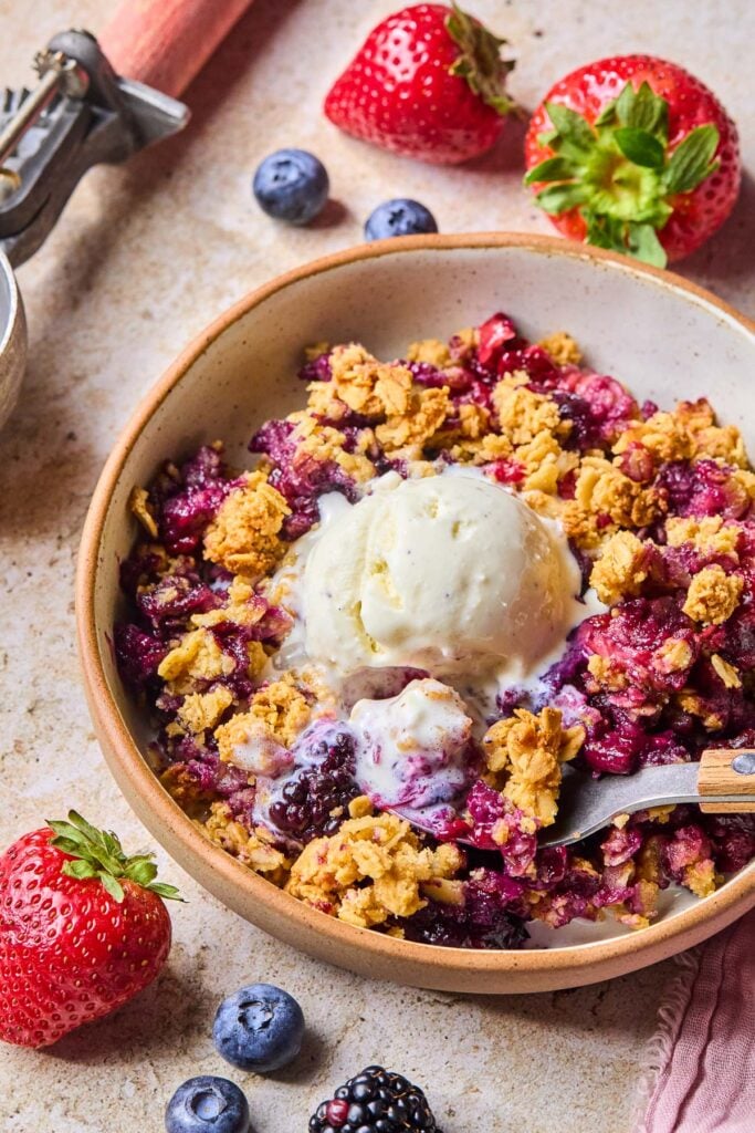 Mixed berry crisp topped with a scoop of vanilla ice cream in a bowl with a spoon in it. Around the bowl are some fresh blueberries and strawberries.