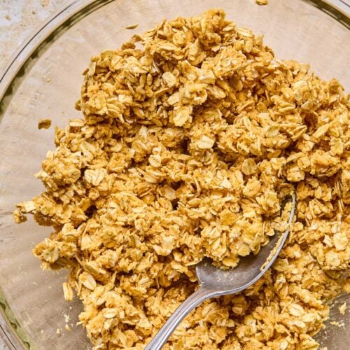 A glass bowl of a mixture of oats, flour, butter, and maple syrup with a spoon in it. Next to the bowl is a small cup of maple syrup.