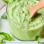 Green goddess dressing in a glass cup with a wooden spoon in it. The cup is on a plate with some fresh herbs on it. Around the plate are more fresh herbs and half a lemon.