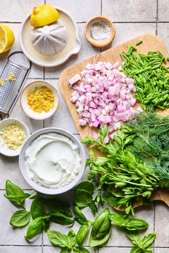 A small bowl of salt and pepper, a wooden cutting board with chopped chives, chopped shallot, fresh dill, and fresh parsley on it, a cup of greek yogurt, some fresh basil leaves, a small bowl of chopped garlic, a small bowl of lemon zest, a small microplane, a half of lemon, and a citrus juicer with a half of lemon on it on a grey tile counter.