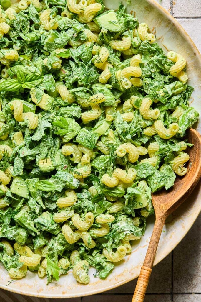 A bowl of green goddess pasta salad with veggies, avocado, and goat cheese with a wooden spoon in it. It is garnished with some fresh herbs.