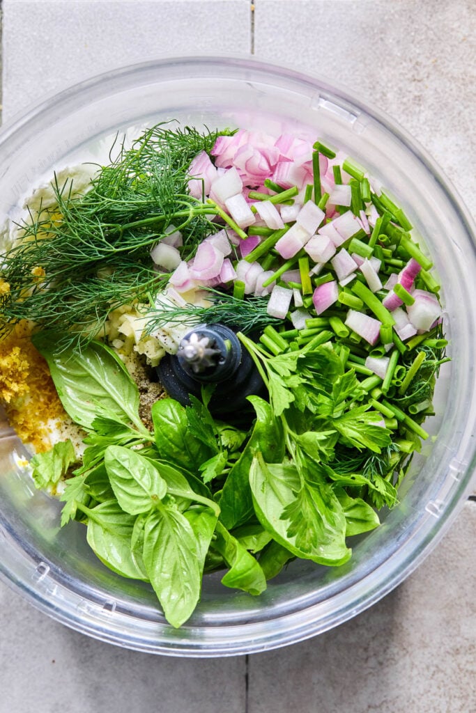 An overhead shot of a food processor bowl filled with fresh basil, greek yogurt, lemon zest, lemon juice, fresh dill, minced garlic, chopped shallot, chopped chives, fresh parsley, salt, and pepper before it is blended.