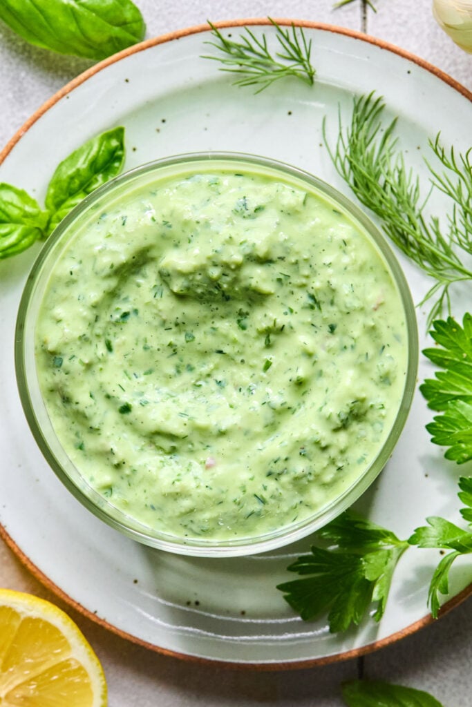 Overhead shot of a glass cup of green goddess dip on a plate with some fresh herbs on it. Around the plate is half a lemon and some more fresh herbs.