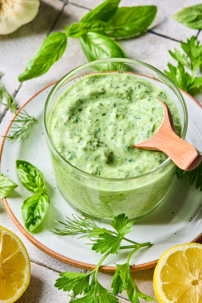 A glass cup of green goddess sauce with a small wooden spoon in it on a plate with some fresh herbs on it. Around the plate are two halves of a lemon, some more fresh herbs, and a bulb of garlic.