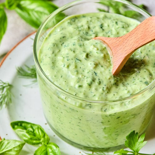 Green goddess dressing in a glass cup with a wooden spoon in it. The cup is on a plate with some fresh herbs on it. Around the plate are more fresh herbs and half a lemon.