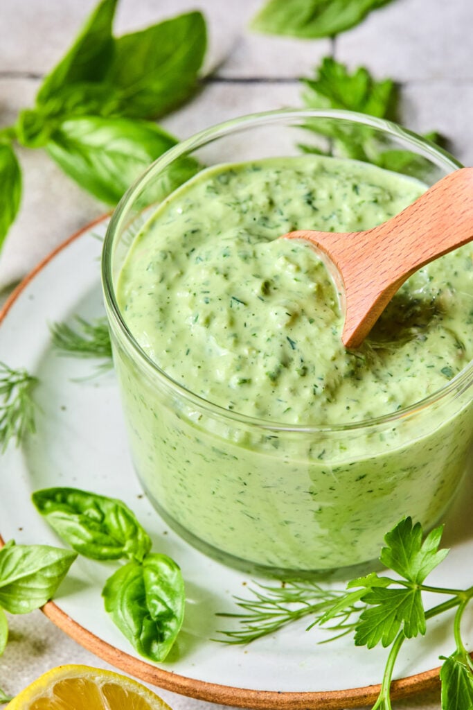 Green goddess dressing in a glass cup with a wooden spoon in it. The cup is on a plate with some fresh herbs on it. Around the plate are more fresh herbs and half a lemon.