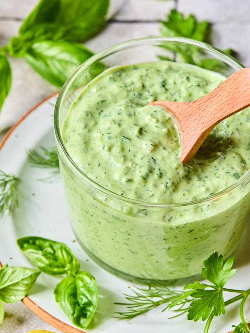 Green goddess dressing in a glass cup with a wooden spoon in it. The cup is on a plate with some fresh herbs on it. Around the plate are more fresh herbs and half a lemon.