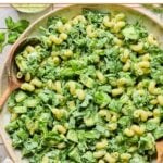 A large serving bowl of green goddess pasta salad topped with fresh herbs, with two wooden spoons in the bowl. Around the bowl are some fresh herbs, a striped kitchen towel, and a cup of green goddess dressing.