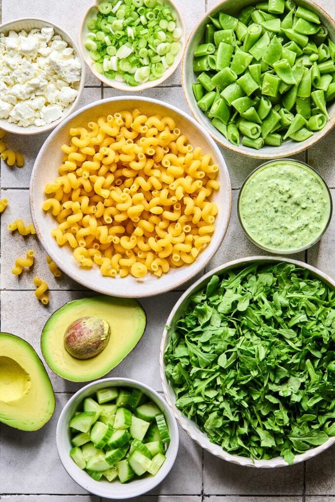 A bowl of chopped sugar snap peas, a cup of green goddess dressing, a bowl of chopped arugula, a bowl of diced cucumber, an avocado that has been cut in half, a bowl of uncooked cavatappi pasta, a bowl of goat cheese, and a bowl of sliced green onions on a grey tile counter.