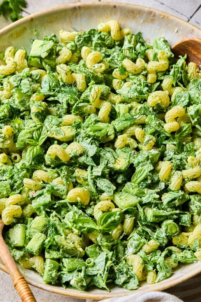 Green goddess pasta with veggies, avocado, goat cheese, and fresh herbs in a bowl. Two wooden serving spoons are in the bowl.