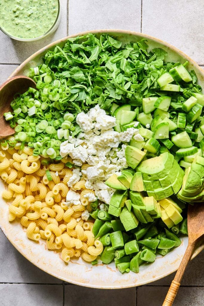 An overhead shot of a large bowl filled with cooked cavatappi pasta, sliced green onions, chopped arugula, diced cucumbers, diced avocado, chopped sugar snap peas, crumbled goat cheese, salt, and pepper before it is mixed. Two wooden spoons are in the bowl, and next to the bowl is a cup of creamy green dressing.