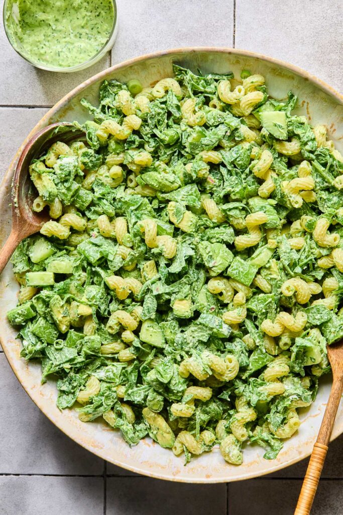An overhead shot of a large bowl filled with pasta, veggies, avocado, and goat cheese tossed with a creamy green dressing. Two wooden spoons are in the bowl, and next to the bowl is a cup of more dressing.