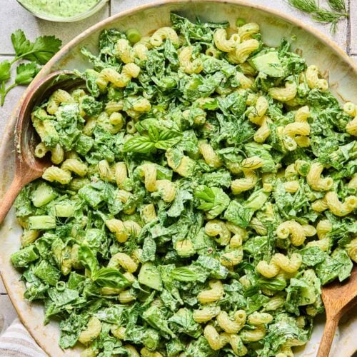 A large serving bowl of green goddess pasta salad topped with fresh herbs, with two wooden spoons in the bowl. Around the bowl are some fresh herbs, a striped kitchen towel, and a cup of green goddess dressing.