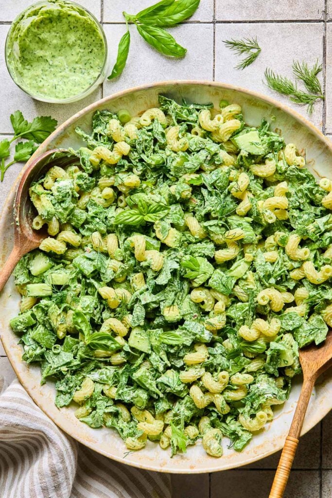 A large serving bowl of green goddess pasta salad topped with fresh herbs, with two wooden spoons in the bowl. Around the bowl are some fresh herbs, a striped kitchen towel, and a cup of green goddess dressing.