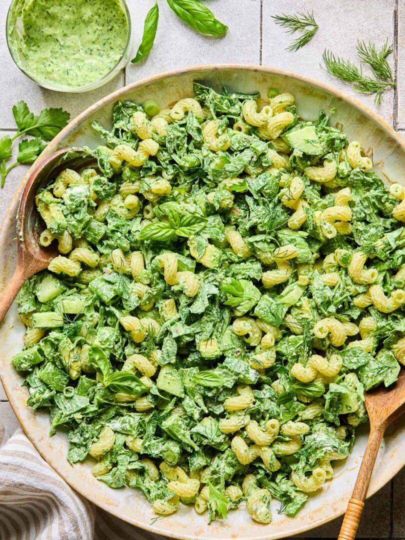 A large serving bowl of green goddess pasta salad topped with fresh herbs, with two wooden spoons in the bowl. Around the bowl are some fresh herbs, a striped kitchen towel, and a cup of green goddess dressing.