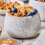 A glass cup with kefir chia pudding topped with granola, blueberries, sliced banana, and cinnamon. Around the cup are a spoon, a bowl of granola, a bowl of blueberries, and another cup of kefir pudding.