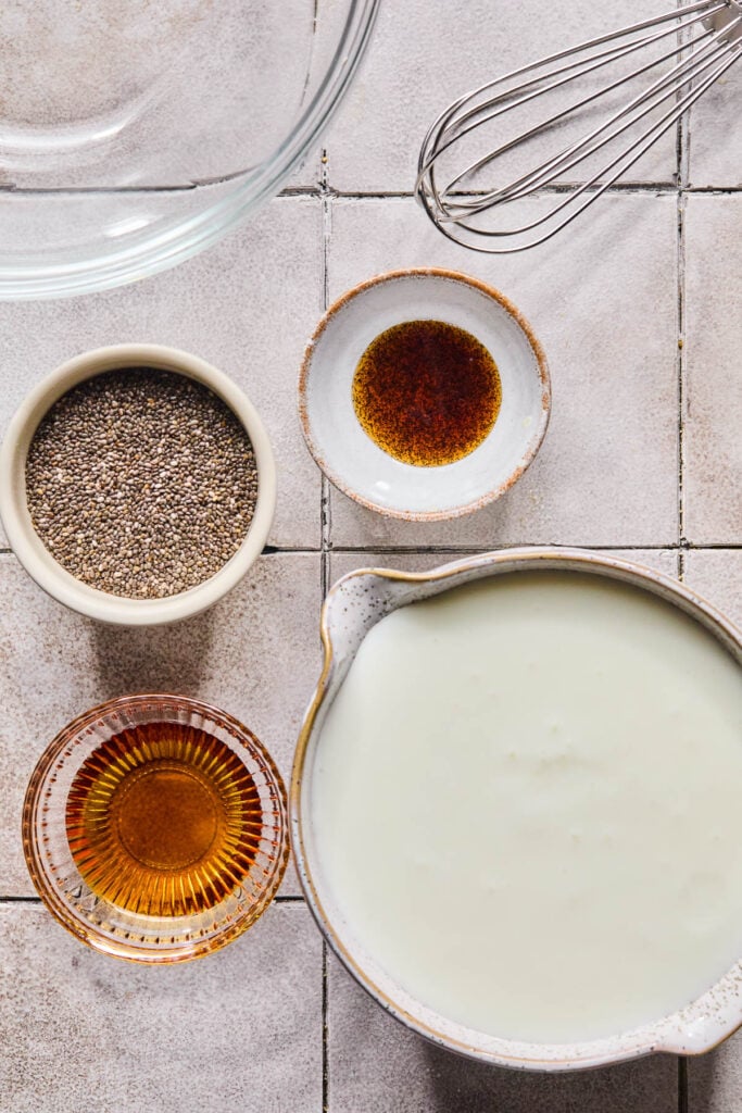 A metal whisk, a small bowl of vanilla bean paste, a bowl of plain kefir, a small bowl of maple syrup, a bowl of chia seeds, and a large glass bowl on a grey tile counter.